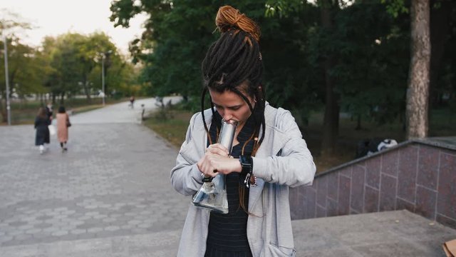 Portrait of young pretty woman with dreadlocks smoking marijuana with a bong in park
