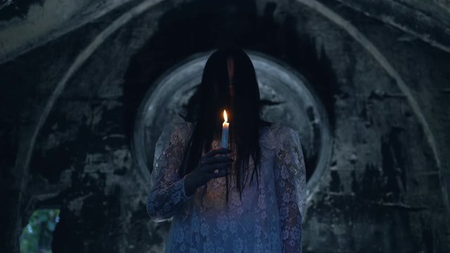 Ominous witch with burning candle near stone crypt, black magic ritual