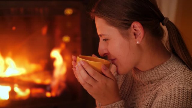 Portrait of smiling young woman looking on the fire in firepalce and drinking hot tea at night