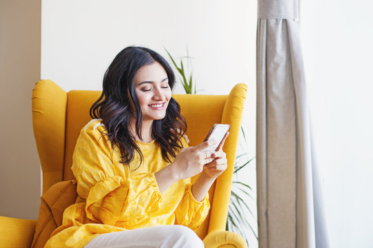 Young Beautiful Indian Woman Using Her Mobile Phone While Sitting In The Armchair At Home