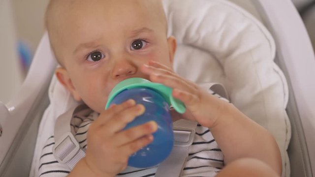 A close-up portrait shot of a lovely little boy lying in baby seat and drinking from non-spill sippy cup