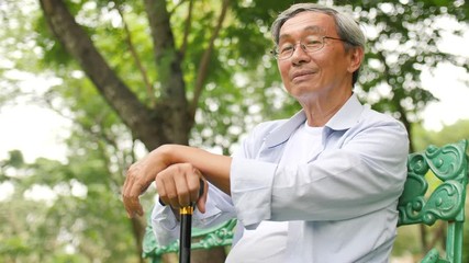 Asian senior man holding a cane and sitting on a bench in the park - Powered by Adobe