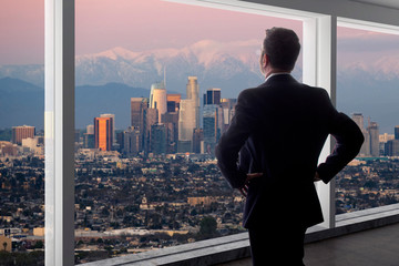 Businessman looking at the buildings of downtown Los Angeles from an office window.  The man looks...