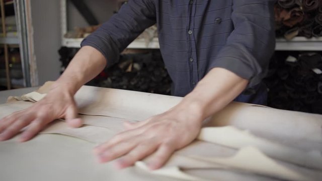 Closeup hands of unrecognizable shoemaker unrolling and examining beige textile standing at table in workshop