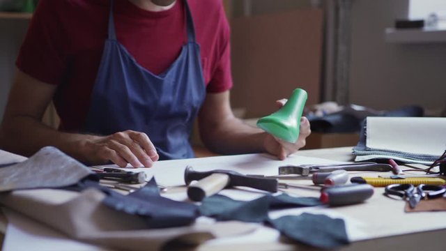Unrecognizable young shoemaker examining shoe last sitting at desk surrounded by tools, textile and leather samples, tracking shot closeup