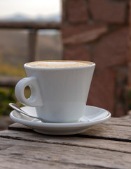 A cappuccino Cup with saucer and spoon sits on an old wooden table outside
