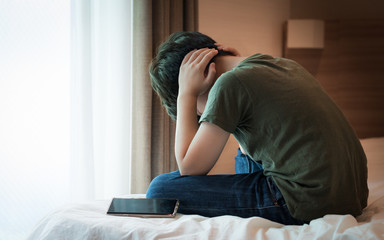 Cyberbullying concept. Young Asian preteen/teenage boy sit cross legged alone next to a smartphone on the bed, covering his ears with hands. Stressed, frustrated, overwhelmed with online bullying.
