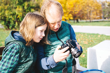 A father and son of school age are talking in close proximity with photo camera sitting on bench. Autumn landscape in yellow tones, warm vests and shirts. Sunny day. Parenthood and childhood concept