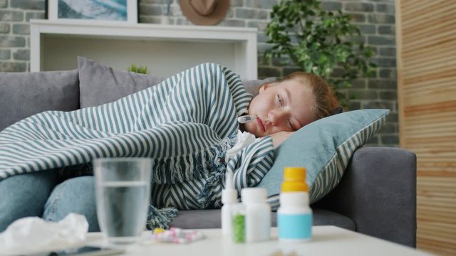 Feverish young woman covered with warm plaid is checking body temperature lying on couch at home feeling bad. Youth, lifestyle and healthcare concept.
