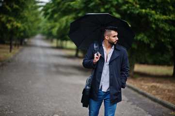 Fashionable tall arab beard man wear on black coat with umbrella and bag case posed at rain weather day.