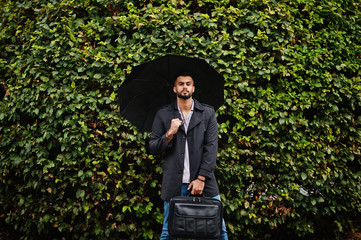 Fashionable tall arab beard man wear on black coat with umbrella and bag case posed at rain weather...