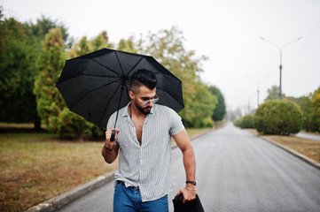 Fashionable tall arab beard man wear on shirt, jeans and sunglasses walking at park with umbrella and coat at hand.