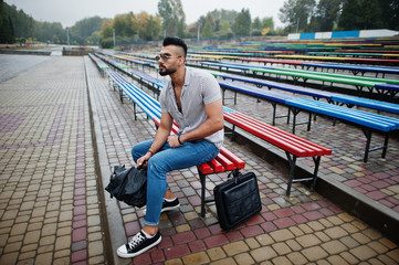 Fashionable tall arab beard man wear on shirt, jeans and sunglasses sitting on colored row of benches.