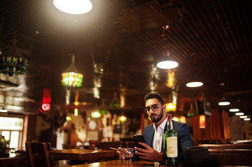 Handsome well-dressed arabian man with glass of whiskey and cigar hold mobile phone,  posed at pub.