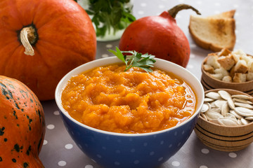 Pumpkin porridge in a bowl among orange pumpkins and seeds. Close-up.