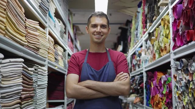 Young shoemaker in apron walking along leather and textile storage towards camera and standing cross-armed looking at camera and smiling, lockdown medium shot