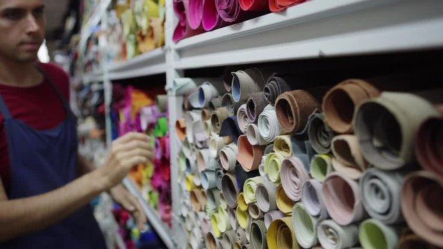 Closeup of young shoemaker choosing shoe material in large storage with multicolored pieces of leather and textile. Craftsman taking roll of blue eco leather from shelf