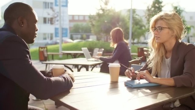 Medium shot of two young business colleagues, Caucasian woman and black man, sitting at table in outdoor cafe or coworking space and discussing work over cup of coffee
