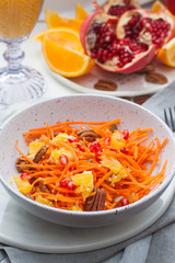 Healthy raw vegetarian salad with carrot, pomegranate seeds, oranges, pumpkin seeds, pecan nuts in grey bowl. Glass of orange juice and pomegranate on the background.
