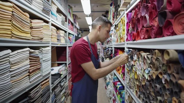 Medium shot of young shoemaker walking along shelves in leather and textile storage and choosing material for his future work. Craftsman taking leather roll from shelf
