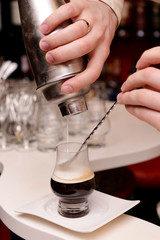 The bartender pours cream from a shaker into a brown cocktail in a glass.

