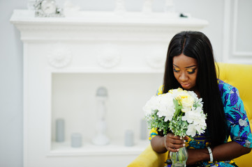 Elegance african american woman model wear in colored clothes. Chic afro women in room sitting on yellow chair with flowers in hand.