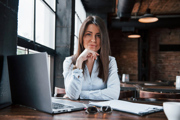 Brunette sitting near the silver colored laptop. Businesswoman in official clothes is indoors in cafe at daytime