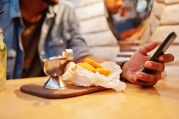 Portrait of handsome successful bearded south asian, young indian freelancer in blue jeans shirt sitting in cafe with chicken nuggets and lemonade. Hold mobile phone at hand.
