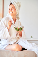 Obraz na płótnie Canvas Picture of happy girl in bathrobe with salad in bowl in her hands sitting on bed.