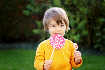 Portrait of little boy with funny hair eating big red lollipop outdoors on green grass looking at camera. Sweet tooth. Carefree childhood. Summer lifestyle