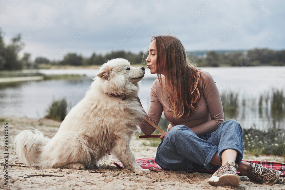 Wall mural Giving a kiss. Blonde girl with her cute white dog have a great time spending on a beach