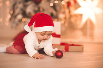 Obraz na płótnie Canvas Funny baby girl 1-2 year old wearing santa claus suit playing with xmas decorations lying on floor in room under Christmas tree closeup. Winter holiday season. Childhood.