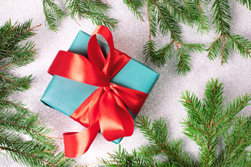 christmas gift box with red ribbon and bow framed by christmas tree branches on silver white glitter background