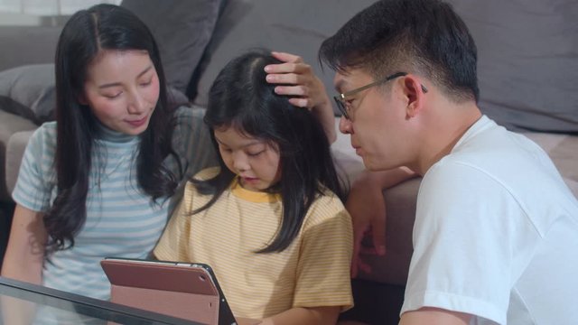 Young Asian family and daughter happy using tablet at home. Japanese mother, father relax with little girl watching movie lying on sofa in living room. Funny parent and lovely child are having fun.