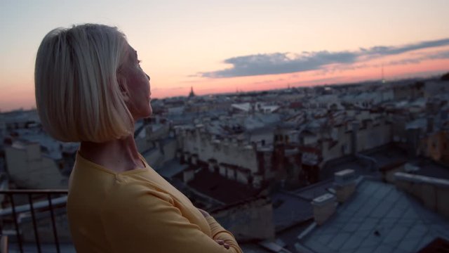 Rear view of blonde middle-aged Caucasian woman wearing casual clothes standing on roof and enjoying cityscape