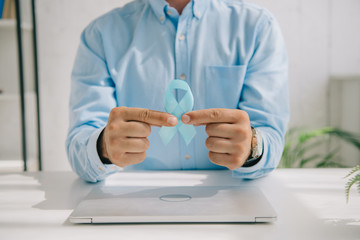 cropped view of man in blue shirt holding blue awareness ribbon near laptop