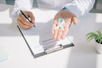 cropped view of doctor holding blue awareness ribbon while writing in medical report