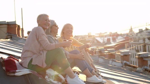 Wide shot of three middle-aged women sitting together on old roof, enjoying cityscape and talking