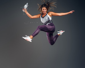 Exited cheerful woman is jumping at studio while holding the bottle of water over grey background.