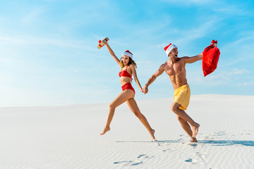 sexy and smiling girlfriend holding gift and boyfriend with santa sack running on beach in Maldives