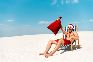 handsome and sexy man in santa hat holding santa sack and sitting on deck chair on beach in Maldives