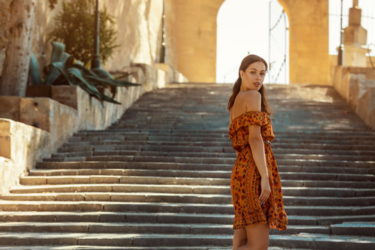 young brunette woman wearing a floral pattern summer dress, walking down a staircase, looking back over her shoulder, at a castle in Artà on Mallorca