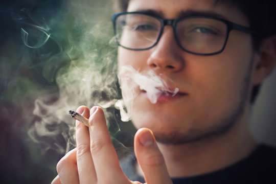 A young handsome guy in glasses smokes a cigarette and exhales smoke, which is illuminated by sunlight, getting untold pleasure from it.