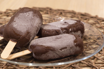 Delicious vegan homemade dessert. Fresh raw popsicles on sticks coated with chocolate glaze lay in glass transparent bowl on wicker tablecloth. Top angle view. Close-up. Selective focus. Healthy meal