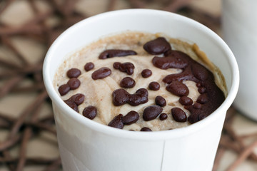 Close up healthy vegan sweet frozen ice cream in a white paper cup. Made from raw banana with coconut cream. Coated with natural chocolate droplets. Table covered with wicker tablecloth