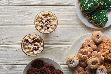 top view of cacao with marshmallow and cacao powder in mugs near Christmas cookies on white wooden table