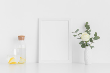 White frame mockup with a rose and eucalyptus in a vase, glass and a bottle on a white table. Portrait orientation.