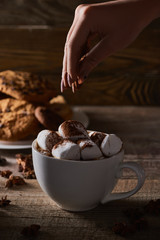 cropped view of woman sprinkling cacao powder on marshmallow in cup on wooden table with anise
