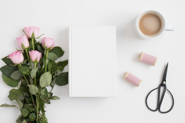 White book mockup with a bouquet of pink roses and workspace accessories on a white table.