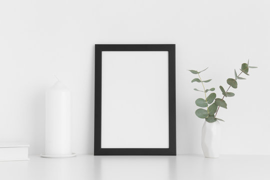 Black frame mockup with workspace accessories and eucalyptus in a vase on a white table. Portrait orientation.